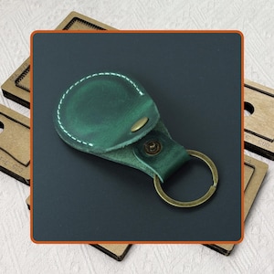 以下が含まれることがあります： 真鍮色のキーリングとボタン留めが付いた、緑色の革製キーホルダー。ホルダーは白い糸でステッチされ、丸みを帯びた上部があります。背景は濃いグレーです。