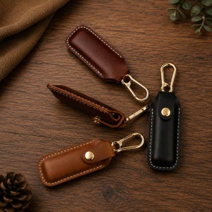 May include: Three leather keychains in brown, black, and tan, each with a gold-colored clasp and button closure. The keychains are rectangular with white stitching and are displayed on a wooden surface.