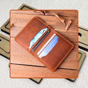 Puede incluir: Un tarjetero de cuero marrón con múltiples ranuras para tarjetas. La cartera está abierta, revelando varias tarjetas de crédito y una tarjeta gris oscuro. La cartera está sobre una superficie de madera.