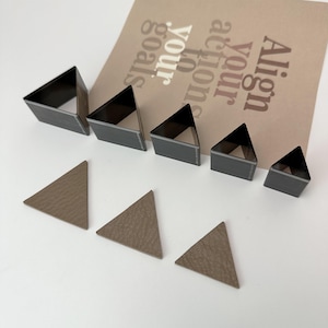May include: Set of six triangle-shaped metal craft punches in various sizes. The punches are arranged in order of size, with the largest punch at the top and the smallest punch at the bottom. The punches are on a white surface. There are also three brown leather triangles cut out with the punches.