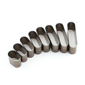 Può includere: Set di sette strumenti ovali per la lavorazione della pelle, ciascuno con un esterno marrone scuro e un interno argentato. Gli strumenti sono disposti in diagonale, con dimensioni crescenti da sinistra a destra. Strumenti per la lavorazione della pelle.