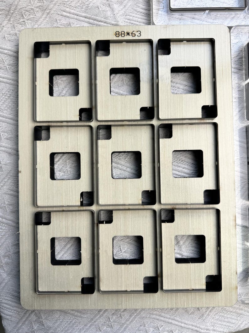 May include: A rectangular metal die featuring nine square apertures, each with a central square cut-out. The die is marked with the numbers "88*63". The metal has a light colour. The dimensions are 88mm by 63mm.