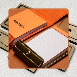 May include: An orange Rhodia notepad with a leather cover, open to a grid-lined page. The cover is brown with white stitching and a dark green leather pen holder. The word "RHODIA" is printed on the notepad.