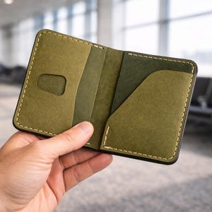 May include: Open olive green leather wallet with multiple card slots and a pocket. The wallet is stitched with white thread and has a black border. The wallet is held open, showcasing its interior.