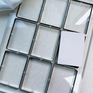 May include: A tray with nine rectangular compartments, each outlined in black. Eight compartments are empty, while one holds a white card. The tray is on a light-coloured surface.