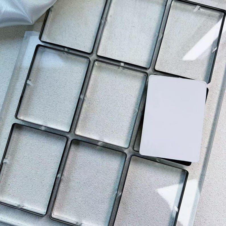 May include: A tray with nine rectangular compartments, each outlined in black. Eight compartments are empty, while one holds a white card. The tray is on a light-coloured surface.