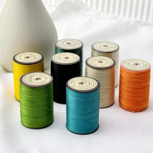 Puede incluir: Una colección de coloridas bobinas de hilo en varios tonos, incluyendo amarillo, verde, negro, turquesa, beige y naranja. Cada bobina tiene una parte superior marrón con una etiqueta circular. Un jarrón blanco en el fondo.