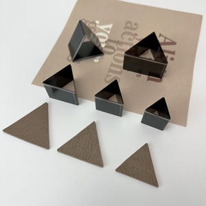 May include: Set of six triangle-shaped metal craft punches in various sizes. The punches are black and have a shiny finish. The punches are arranged on a brown paper background with the text "All our actions speak volumes".