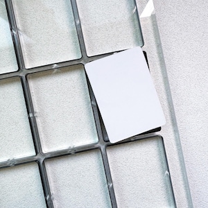 Puede incluir: Una paleta rectangular transparente con múltiples compartimentos cuadrados, cada uno delineado en negro. Una tarjeta blanca está parcialmente insertada en uno de los compartimentos, lo que sugiere una herramienta de exhibición u organización de productos. La paleta se encuentra sobre una superficie clara texturizada.