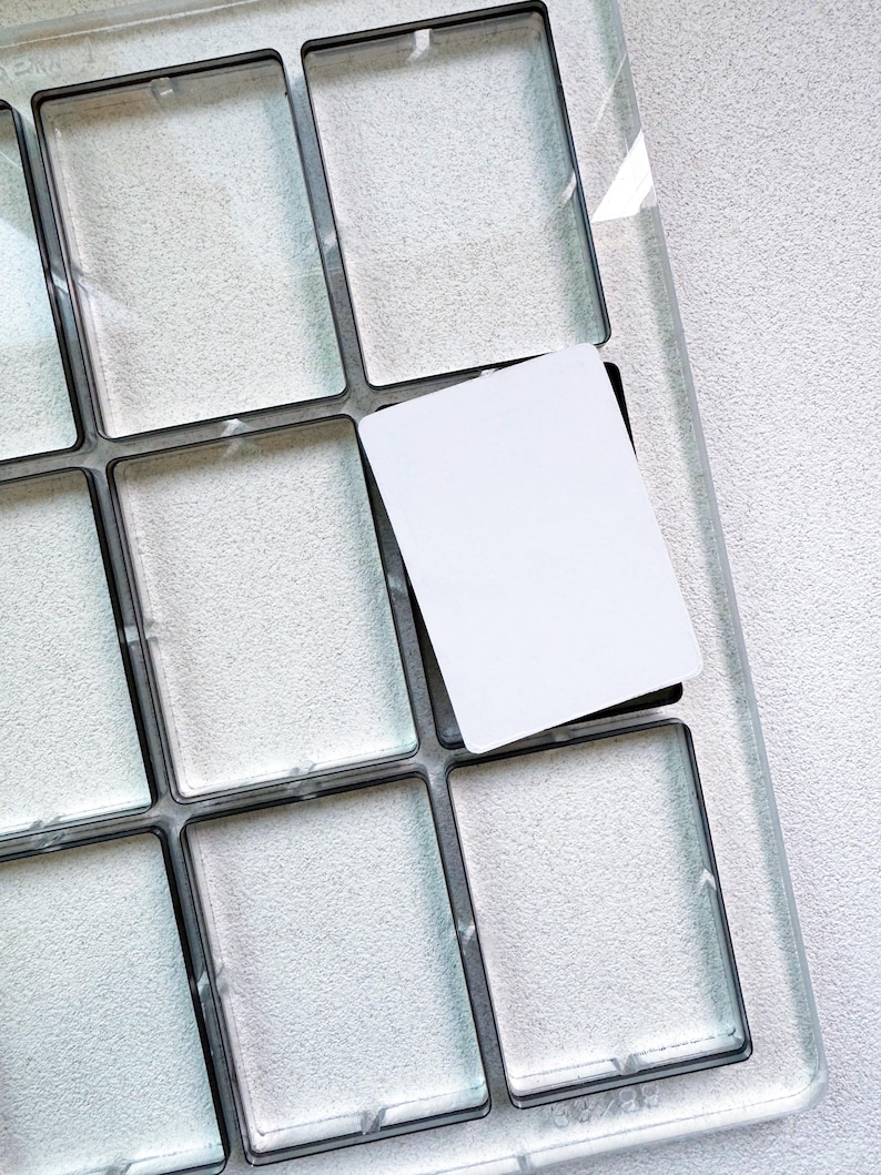 May include: A clear, rectangular palette with multiple square compartments, each outlined in black. A white card is partially inserted into one of the compartments, suggesting a product display or organization tool. The palette sits on a textured, light-coloured surface.