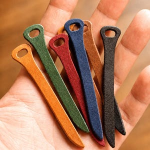 Könnte beinhalten: Eine Sammlung farbenfroher Leder-Kabelhalter in Orange, Grün, Rot, Blau, Braun und Schwarz. Jeder Halter hat oben ein kleines Loch zum Befestigen an Kabeln oder anderen Gegenständen.