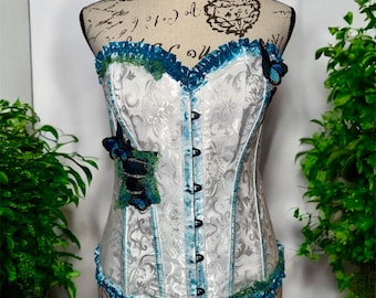 May include: A white corset with a blue and green lace trim and butterfly embellishments. The corset has a busk closure in the front and is made of a satin-like fabric.