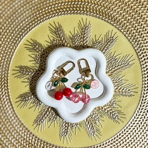 Puede incluir: Dos llaveros con forma de cereza con herrajes dorados descansan sobre un plato blanco con forma de flor. Un llavero tiene cerezas rojas, el otro tiene cerezas rosas. Cada uno tiene hojas verdes y un pequeño dije con una letra.
