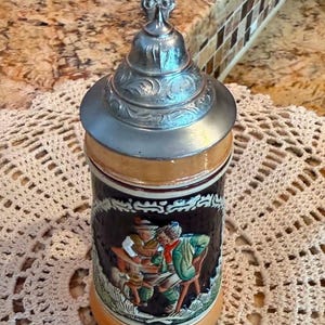 May include: A ceramic beer stein featuring a dark brown body with a relief scene of two figures sitting at a table. The stein is topped with a metallic, ornate, conical lid that includes a decorative finial. The base and rim are light tan.