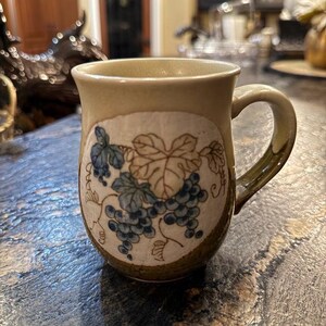 May include: A beige ceramic mug with a brown and blue grape design. The mug has a rounded body and a handle.