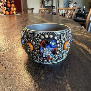 May include: A wide, silver-toned cuff bracelet with colorful, mosaic-style glass accents. The bracelet features a detailed, textured design with small, round embellishments and floral-like patterns. The glass pieces are primarily blue, orange, and red.