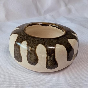 May include: A ceramic bowl with a circular shape and a wide opening. The bowl is off-white with dark brown, dripped-paint-like patterns. The interior is a light beige color. The bowl is likely handmade and suitable for decorative use.