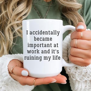 Op de afbeelding: Witte keramische mok met de tekst "I accidentally became important at work and it's ruining my life" in het zwart. De mok heeft een groot handvat en wordt vastgehouden door een persoon die een groen shirt en een witte trui draagt.