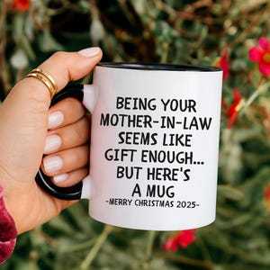 Op de afbeelding: Een witte keramische mok met een zwarte handgreep en rand, vastgehouden door een hand. De mok heeft zwarte tekst die luidt: "JE SCHOONMOEDER ZIJN LIJKT GENOEG CADEAU... MAAR HIER IS EEN MOK - VROLIJK KERSTFEEST 2025-".