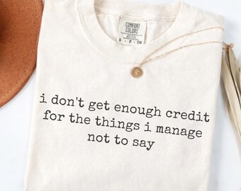 May include: Cream-coloured Comfort Colors t-shirt with the text "i don't get enough credit for the things i manage not to say" in black. A gold necklace with a pendant is draped on the shirt.