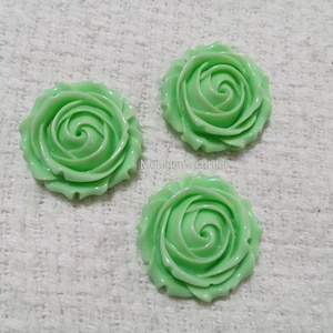 May include: Three mint green rose-shaped cabochons with a smooth, glossy finish and layered petal details. The text "Mangen's corner" is printed in small, light-colored letters between the top two rose-shaped embellishments.