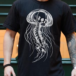 May include: Black t-shirt featuring a white jellyfish design with a skull incorporated into the jellyfish's head. The jellyfish has long, flowing tentacles. The shirt is short-sleeved and made of a soft material.