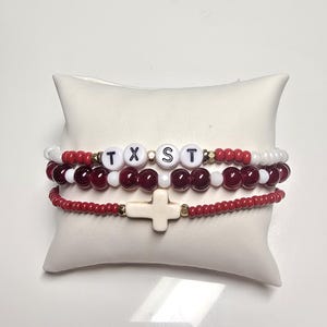 May include: Three beaded bracelets with red, white, and gold beads. One bracelet spells out "TXST" with white letter beads. The bottom bracelet features a white cross charm. The bracelets are displayed on a white pillow.