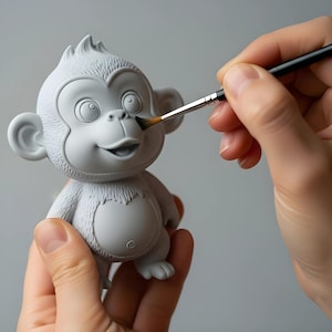 Può includere: Una statuetta di scimmia grigia in stile cartone animato viene dipinta con un piccolo pennello. La statuetta è tenuta tra le mani di una persona e il pennello applica la vernice sul viso della scimmia. La scimmia ha un'espressione sorridente.