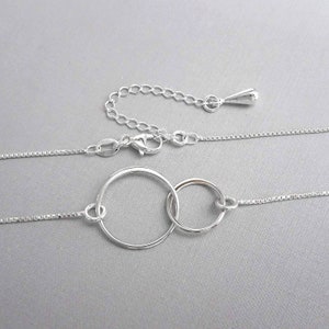 May include: A silver necklace with two interlocking circles as a pendant. The necklace has a delicate chain and a lobster clasp.