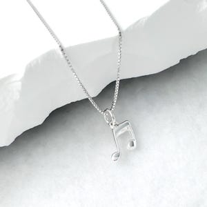 May include: A silver necklace featuring a musical note pendant. The pendant is an eighth note design. The chain is a fine box chain. The background is a white, textured surface, providing a clean contrast to the necklace.