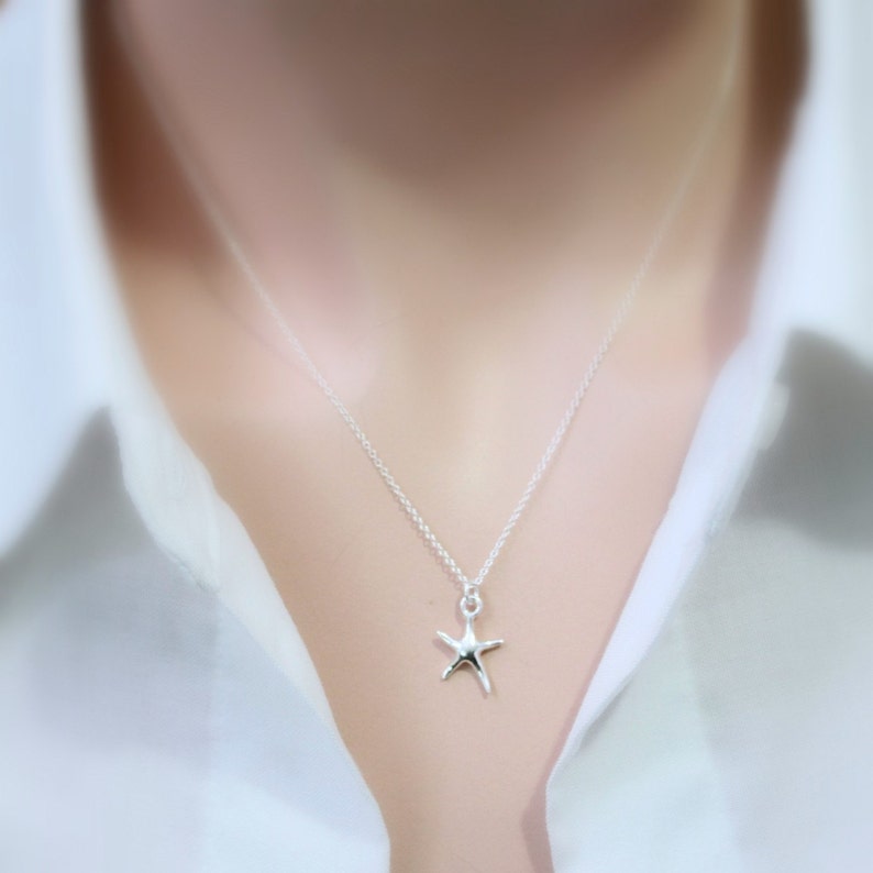 May include: A silver starfish pendant necklace. The necklace features a delicate chain and a small, detailed starfish charm. The jewelry is worn against a white shirt, creating a simple and elegant look. The starfish charm is approximately 0.75 inches in size.