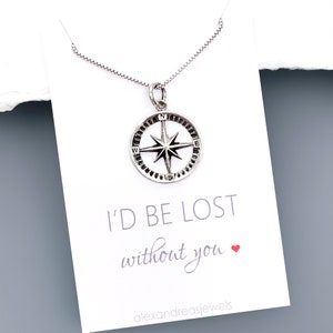 May include: A silver-colored compass pendant necklace on a white card. The compass pendant has a star design in the center and is attached to a delicate chain. The card reads "I'D BE LOST without you" with a small red heart.