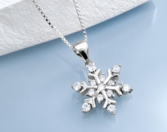 May include: A silver-toned necklace featuring a snowflake-shaped pendant. The pendant is encrusted with multiple clear, round-cut stones that catch the light. The chain is a delicate box-link style, connecting to a polished metal bail at the top.