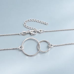 May include: A silver-toned necklace featuring a delicate box chain and two interlocking circular pendants of different sizes. The necklace includes a lobster claw clasp and an adjustable extender chain with small circular links at the end.