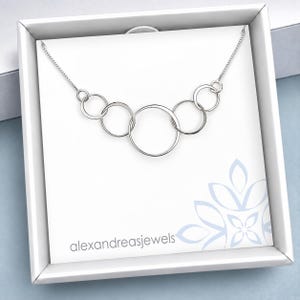 May include: A silver-toned necklace featuring five interconnected circular rings of varying sizes arranged in a horizontal row. The necklace is displayed on a white card inside a square box, with the text alexandreasjewels printed in the bottom left corner.