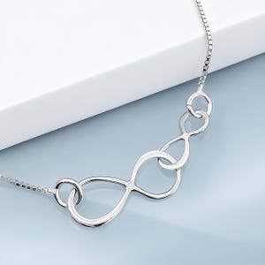 Peut inclure: Un collier argenté doté d'un pendentif central composé de deux symboles infinis entrelacés. Le pendentif est relié à une fine chaîne de type boîte qui s'étend des deux côtés, présentant une finition métallique polie et un design en boucle.