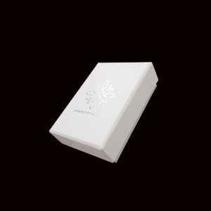 May include: A white rectangular box with a silver floral design and the text "alexandreasjewels" printed on the top. The box is set against a black background.