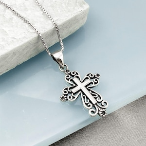 May include: A silver-colored pendant necklace featuring a detailed cross design. The cross has an ornate, openwork pattern with a smaller cross in the center. The necklace chain is a delicate box chain style.