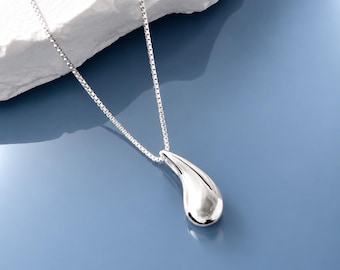 May include: A polished silver-toned necklace featuring a smooth, teardrop-shaped pendant suspended from a delicate box chain. The metallic surface of the pendant reflects light, creating bright highlights against its curved, organic form.