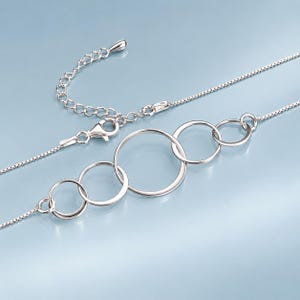 May include: A silver-toned necklace featuring a delicate box chain and a central pendant composed of four interlocking circular rings of varying sizes. The necklace includes a lobster claw clasp and an adjustable extender chain with a small teardrop charm.