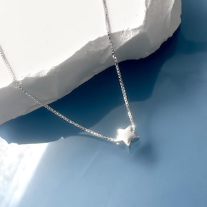May include: A silver chain necklace with a small, star-shaped pendant. The necklace is on a white rock surface with a blue background.