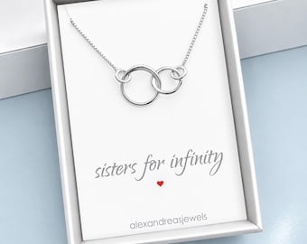 Pode incluir: Um colar de tom prateado com dois pingentes circulares entrelaçados numa corrente fina, apresentado numa caixa branca com o texto "sisters for infinity" impresso em cinzento, um pequeno ícone de coração vermelho e o nome da marca alexandreasjewels.