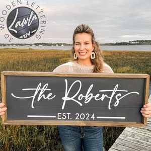 May include: A woman holds a dark gray wooden sign with white lettering that reads "The Roberts EST. 2024".