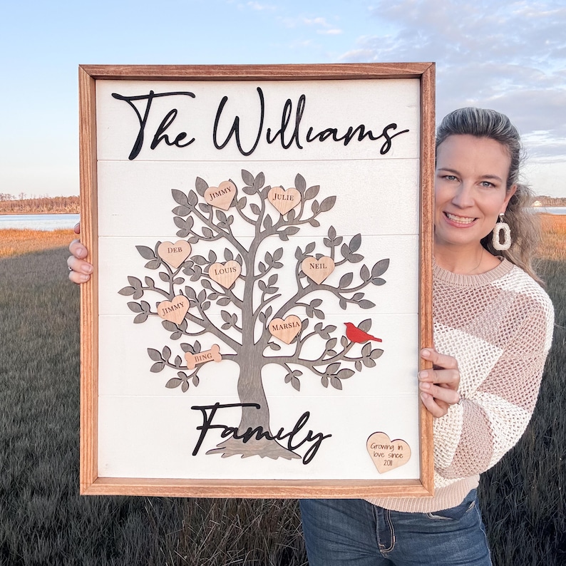 May include: A white wooden sign with a brown frame features a gray tree with brown hearts and a red cardinal. The hearts have names written on them, including "The Williams", "Jimmy", "Julie", "Deb", "Louis", "Marsha", "Neil", and "Bing". The sign also includes the text "Family" and "Growing in love since 2018".