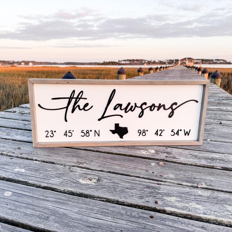 K&ouml;nnte beinhalten: Ein wei&szlig;es Holzschild mit dem Text "The Lawsons" in schwarzer Kursivschrift. Unter dem Text befindet sich eine schwarze Silhouette des Bundesstaates Texas und die Koordinaten "23&deg; 45' 58" N 98&deg; 42' 54" W".