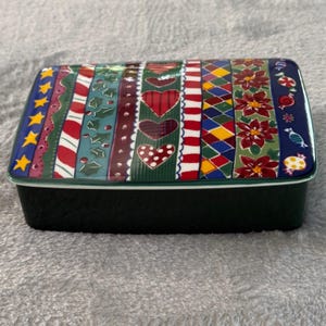 Puede incluir: Una caja rectangular de cerámica para baratijas con una base verde oscuro y una tapa decorativa. La tapa presenta un colorido patrón de rayas con corazones, estrellas y flores de Pascua. La caja probablemente se utiliza para guardar objetos pequeños.