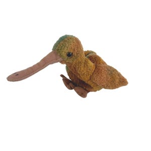 May include: A soft toy kiwi bird in shades of brown, green, and tan. The bird has a long, curved beak and small, dark eyes. The toy is made of a soft, textured material.