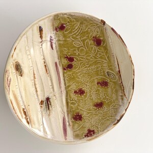 May include: A handmade ceramic plate with a unique design. The plate features a cream-colored birch bark pattern on one side and a green floral pattern with red accents on the other. The plate's rim has a brown border.