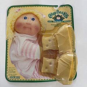May include: A Cabbage Patch Kids doll with blonde hair and blue eyes, wearing a pink and white striped outfit. The packaging includes a pair of yellow doll sneakers with laces. The packaging reads "DOLL SNEAKERS & SOCKS".