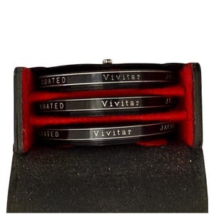 May include: A black leather case with three Vivitar camera lens filters. The filters are black with silver text that reads "Vivitar" and "Coated". The interior of the case is red.